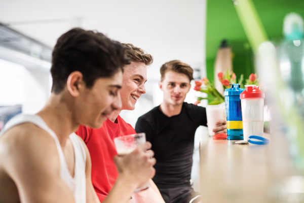 Three young fit men in crossfit gym sitting and holding glasses with protein drinks.