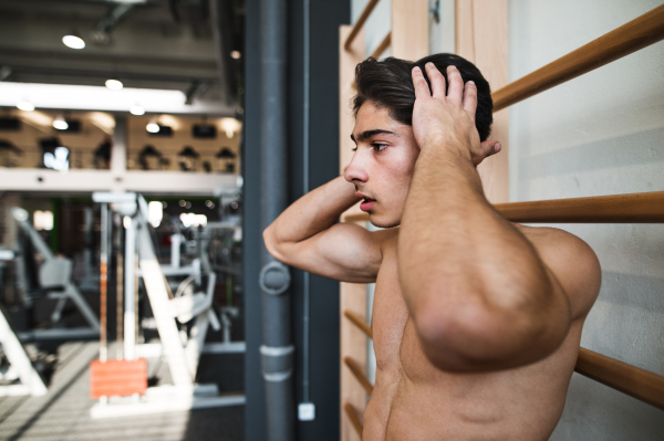 Fit handsome young man in gym standing topless in front of wall-bars.