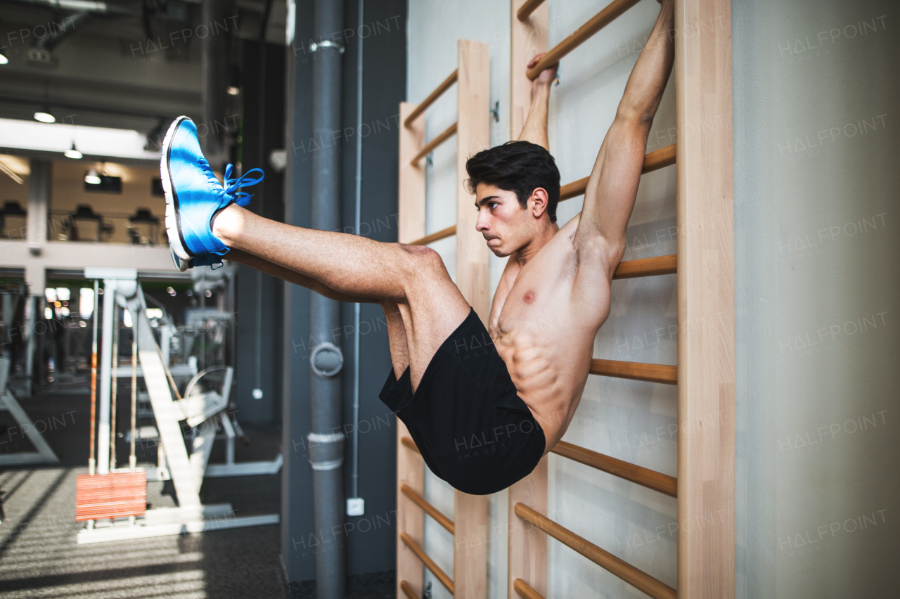 Young handsome fit man in gym working out on wall bars, holding L-sit.