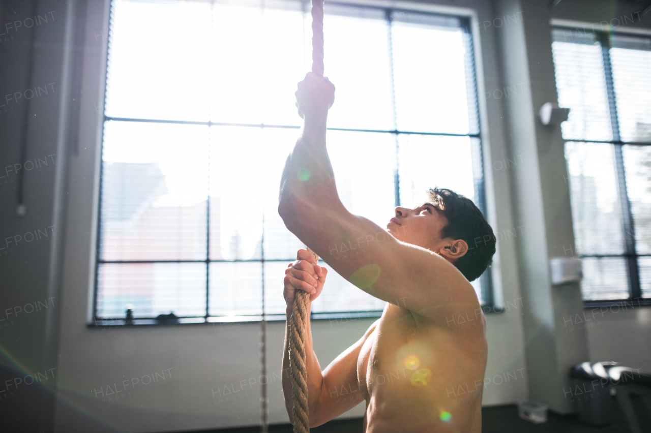 A fit young man in gym standing topless , holding a climbing rope. Copy space.