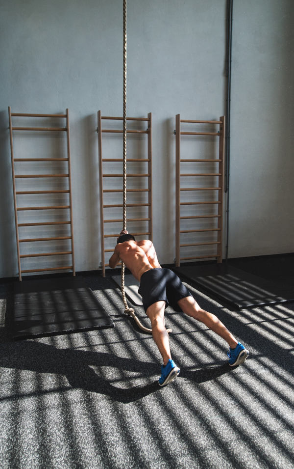 Young fit man in gym working out with climbing rope. Rear view.