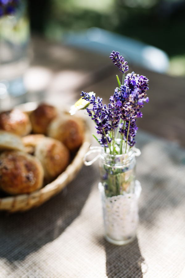 A close-up of a vase with lavender flowers and a basket with bread buns on the table set for a garden party or family celebration outside.