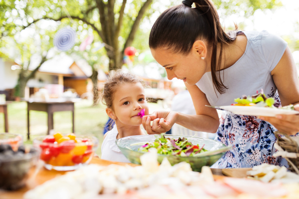 Family celebration outside in the backyard. Big garden party. Birthday party. Young mother with a small girl standing at the table with food.