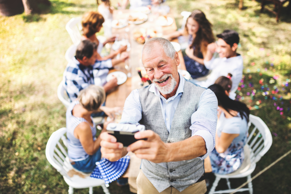 Family celebration outside in the backyard. Big garden party. A senior man taking selfie with a camera.