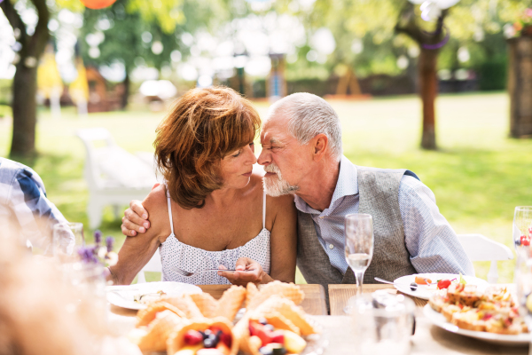 Senior couple sitting at the table on a garden party, touching noses. Family celebration outside in the backyard.