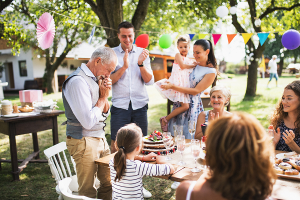 A senior man with an extended family looking at the birthday cake, crying in happiness. Celebration outside in the backyard or big garden party.