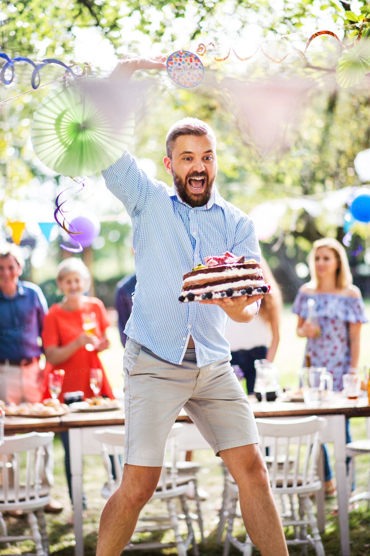 Family celebration outside in the backyard. Big garden or birthday party. Mature man holding and jumping with a birthday cake.