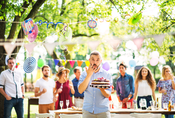 Family celebration outside in the backyard. Big garden or birthday party. Mature man holding a birthday cake and licking his finger.