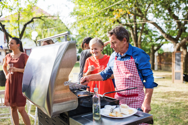 Family celebration outside in the backyard. Barbecue party. An old man cooking food on the grill.