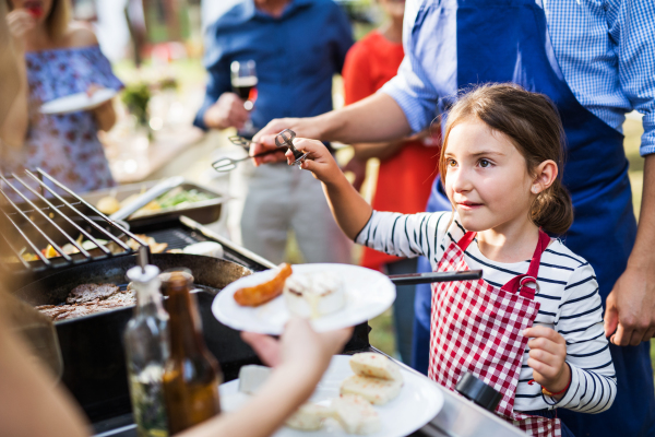 Family celebration outside in the backyard. Barbecue party. An unrecognizable man with a small girl cooking food on a grill.