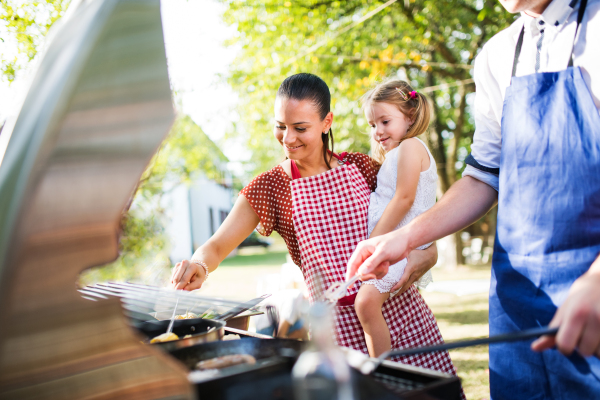 Family celebration outside in the backyard. Barbecue party. A woman holding a small girl and an unrecognizable man cooking.