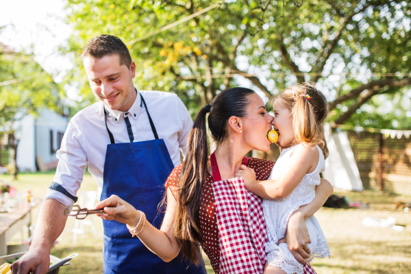 Family celebration outside in the backyard. Barbecue party. A man cooking, a woman holding a small girl and eating corn together.