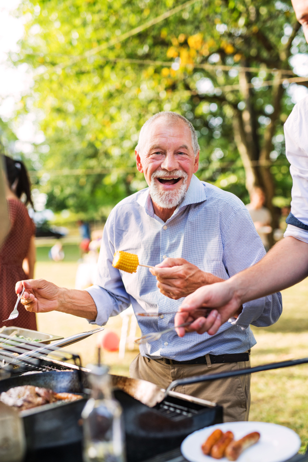 Family celebration outside in the backyard. A happy senior man standing by the grill, holding corn on a barbecue party.