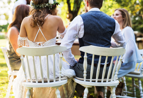 Wedding reception outside in the backyard. Bride, groom and the guests sitting at the table. Rear view.