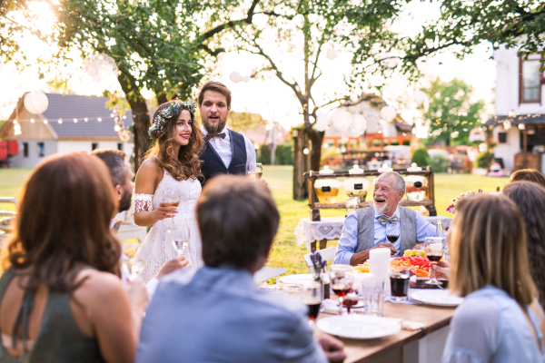 Wedding reception outside in the backyard. Handsome groom giving speech. Guests sitting around the table.