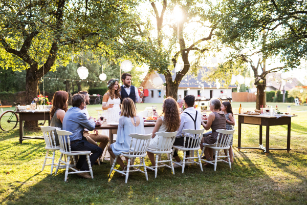 Wedding reception outside in the backyard. Handsome groom giving speech. Guests sitting around the table.