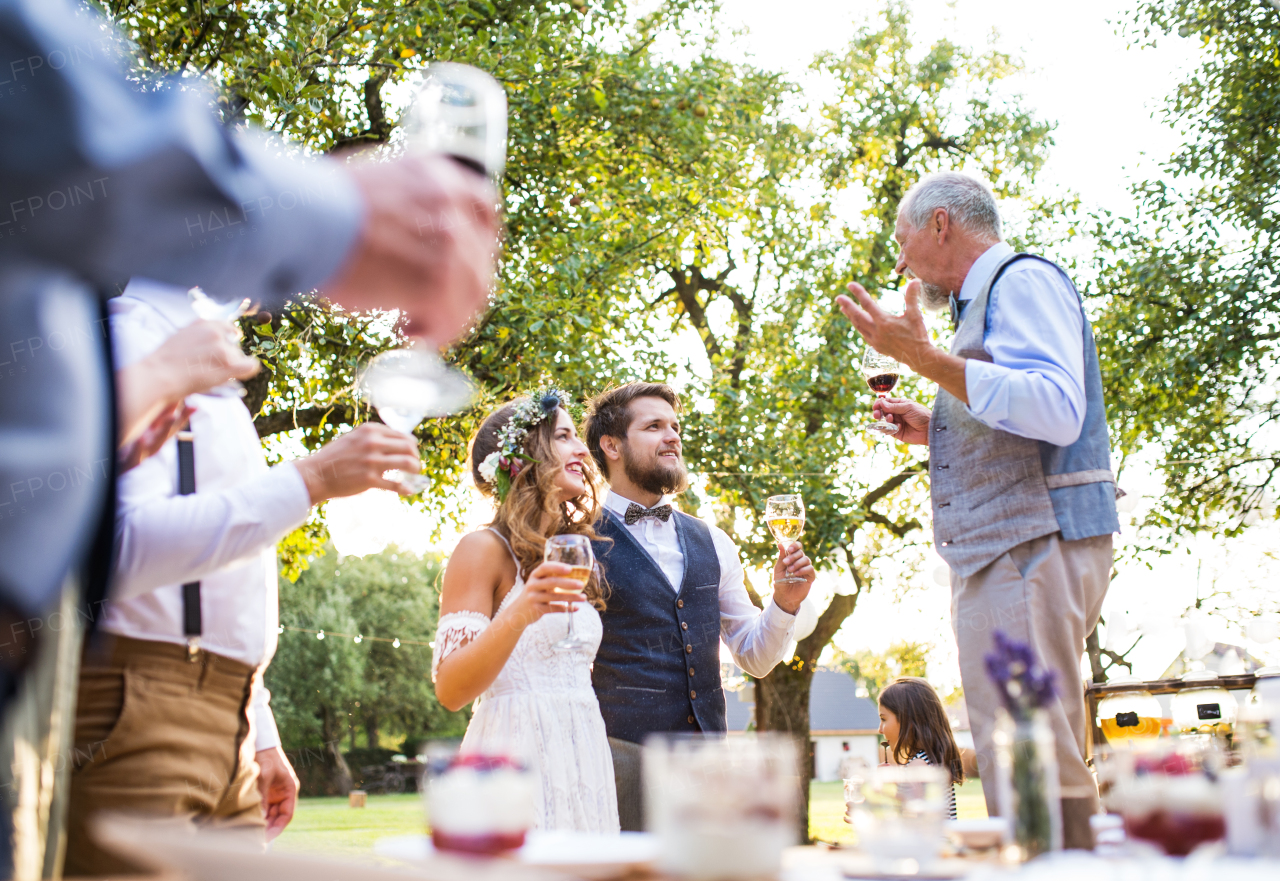 A senior man making speech at wedding reception outside in the background. Bride and groom and other guests holding glasses.