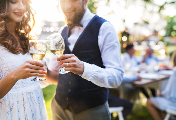 Wedding reception outside in the backyard. Bride and groom clinking glasses with champagne, unrecognizable guests in the background.