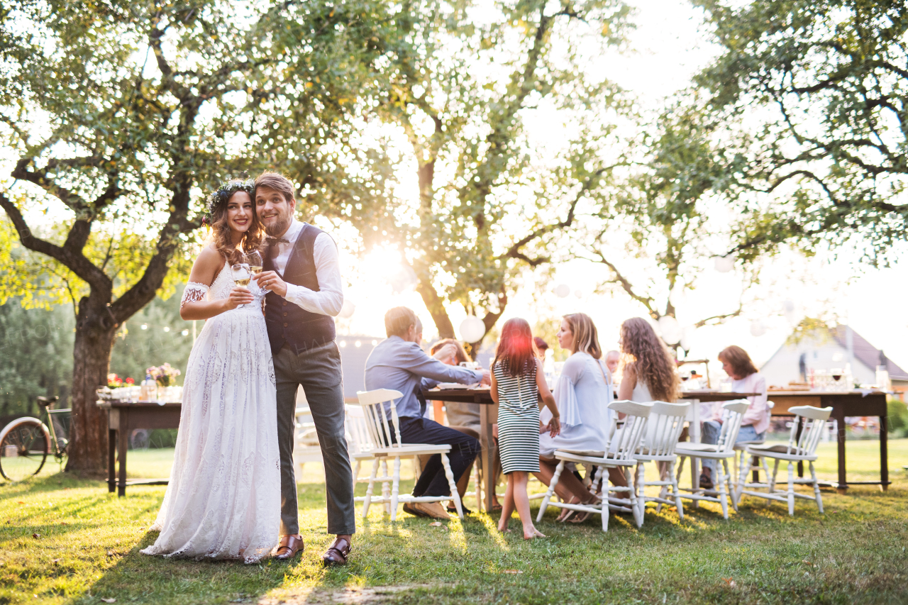 Wedding reception outside in the backyard. Bride and groom clinking glasses with champagne, guests sitting at the table in the background.