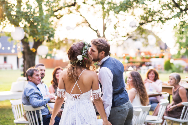 Wedding reception outside in the backyard. Bride and groom kissing, guests sitting at the table in the background. Rear view.