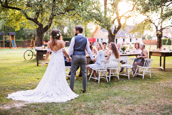 Wedding reception outside in the backyard. Bride and groom holding hands, guests sitting at the table in the background. Rear view.