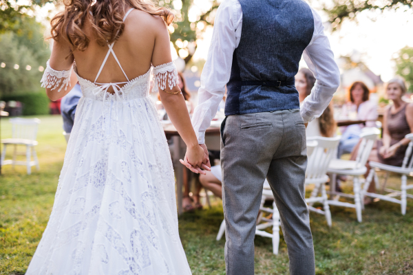 Unrecognizable bride, groom and the guests at wedding reception outside in the backyard, holding hands. Rear view.