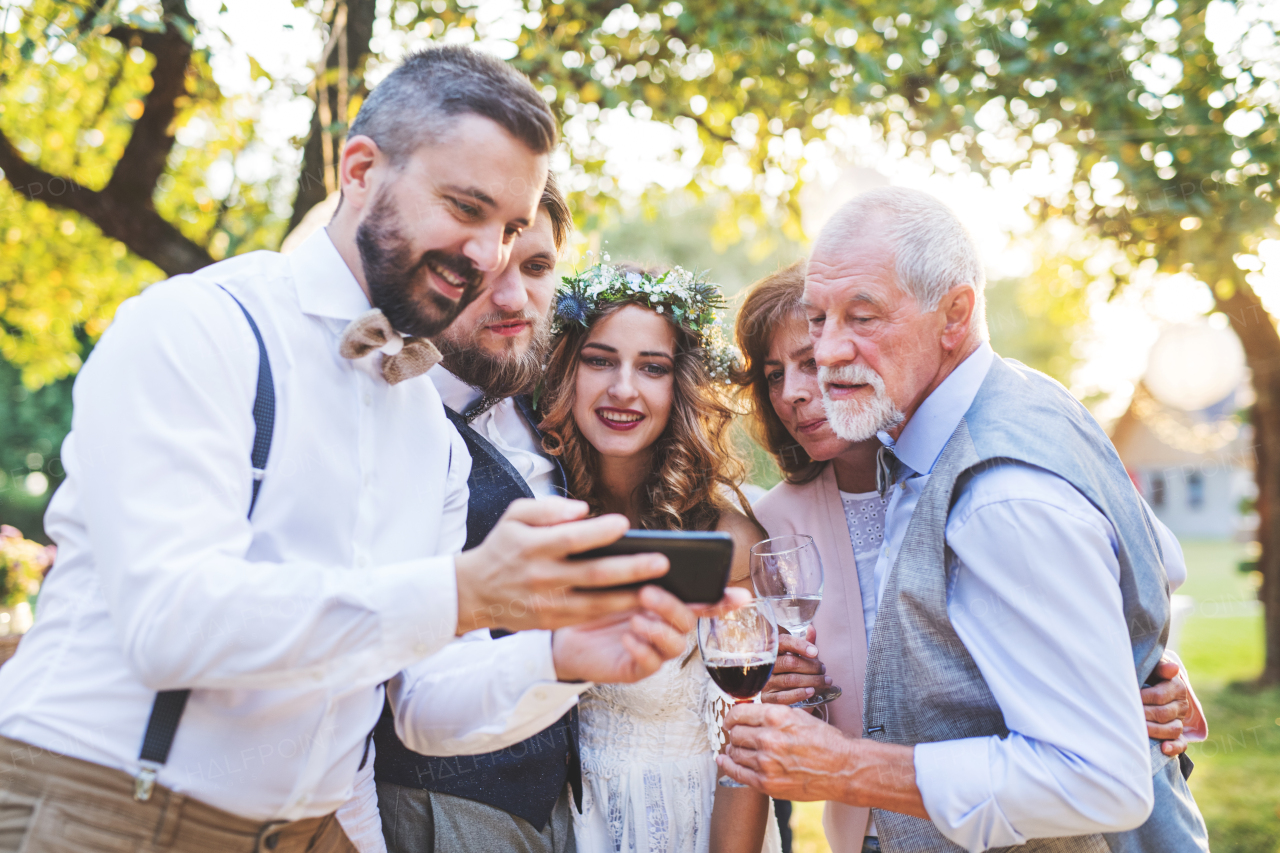 Happy bride, groom and guests with smartphones taking selfie outside at wedding reception.