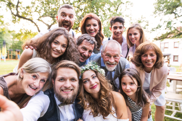 A close-up of happy bride, groom with guests taking selfie at wedding reception outside in the backyard.