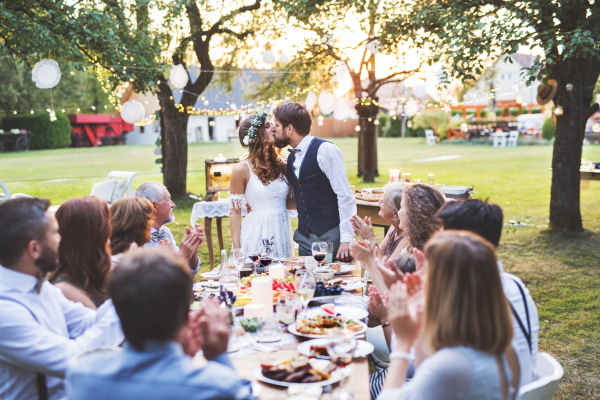 Wedding reception outside in the backyard. Bride and groom kissing each other. Family and guests around the table, clapping.