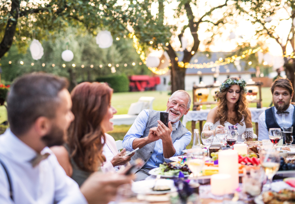 A happy senior man taking selfie at the wedding reception outside in the backyard. Bride, groom and guests sitting at the table.