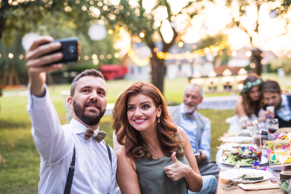 A couple taking selfie at the wedding reception outside in the backyard. Bride, groom and guests sitting at the table.