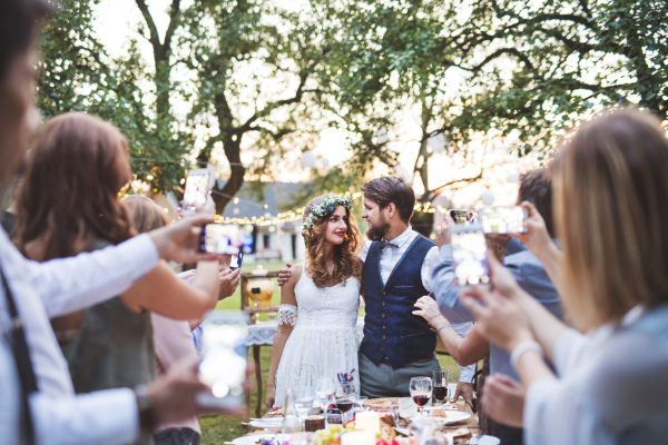 Guests with smartphones taking photo of bride and groom at wedding reception outside in the backyard.