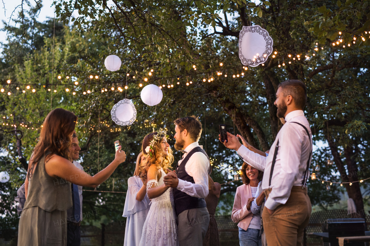 Guests with smartphones taking photo of a dancing bride and groom at wedding reception outside. An evening in the backyard.