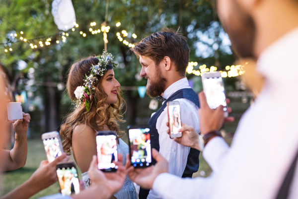 Guests with smartphones taking photo of a dancing bride and groom at wedding reception outside. An evening in the backyard.