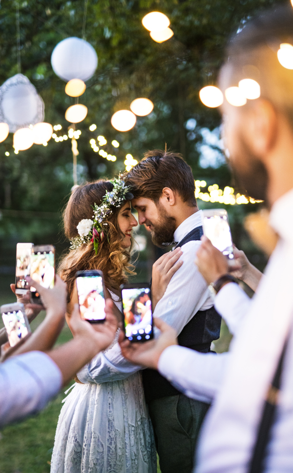 Guests with smartphones taking photo of a dancing bride and groom at wedding reception outside. An evening in the backyard.