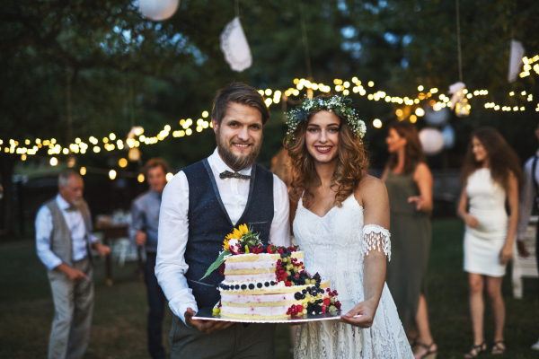 Wedding reception outside in the backyard. Bride and groom holding a cake, guests in the background.