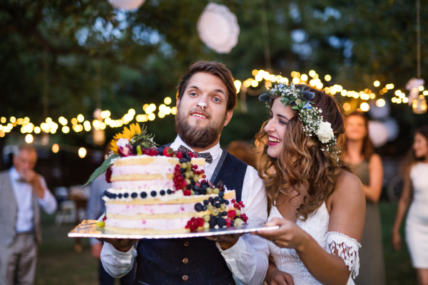 Wedding reception outside in the backyard. Bride and groom holding a cake, guests in the background.