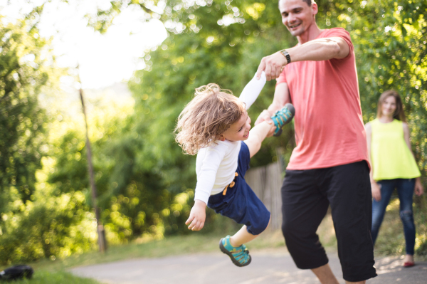 Young father spinning his cute little son. Mother watching them. Sunny summer day in nature.