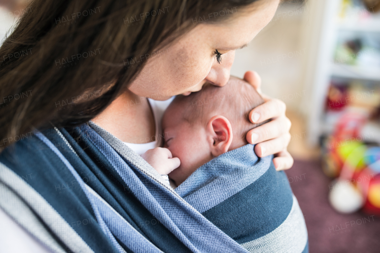 Close up of unrecognizable young mother with her newborn baby son in sling at home, kissing him