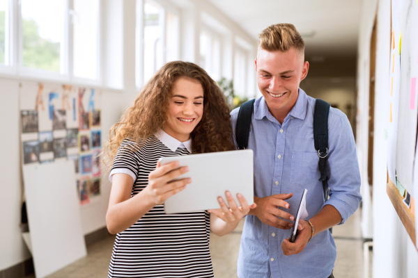 Attractive teenage student couple in high school hall with tablet, taking selfie.