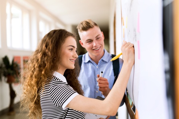 Attractive teenage student couple in high school hall writing something on notice board.