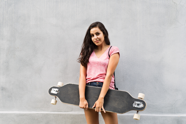 Beautiful teenage student with skateboard standing against concrete wall.