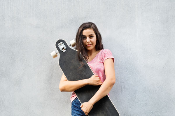 Beautiful teenage student with skateboard standing against concrete wall.