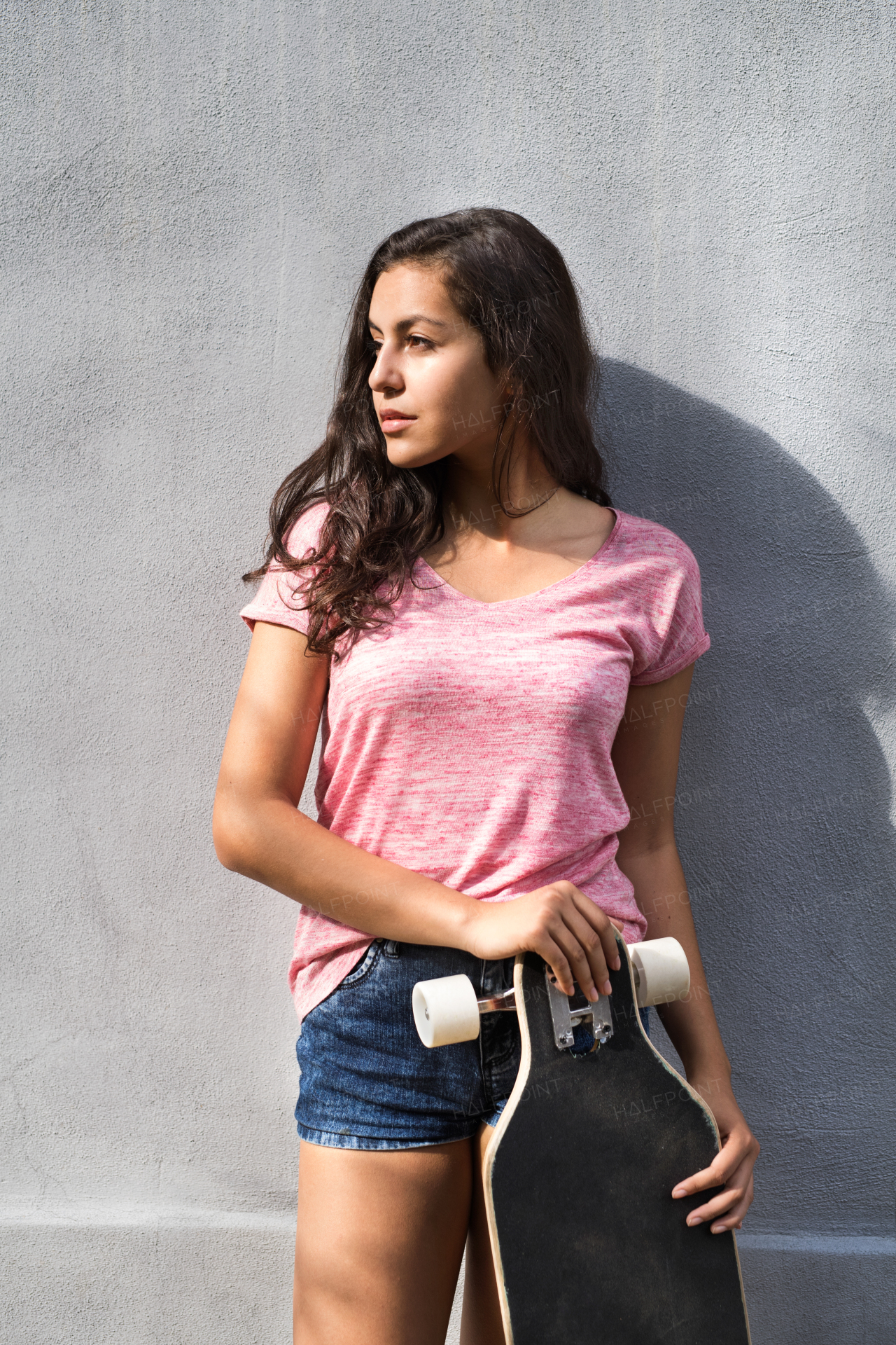 Beautiful teenage student with skateboard standing against concrete wall.