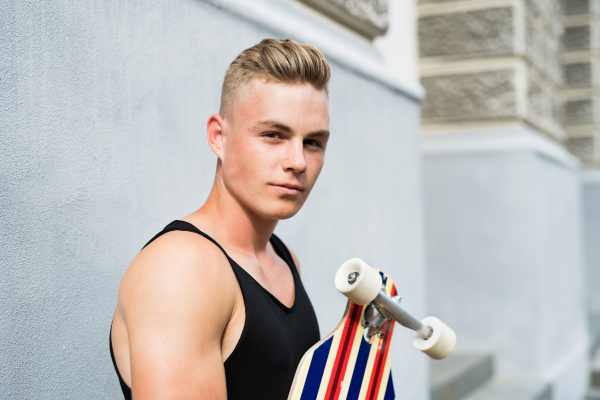 Cool blonde fit teenage boy in black vest holding a scateboard in front of old building.