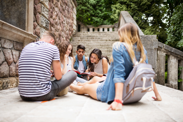 Group of attractive teenage students sitting on stone steps in front of university, reading and studying.