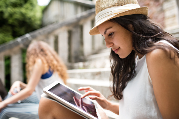 Attractive teenage student sitting on stone steps in front of university holding tablet, reading or watching something.