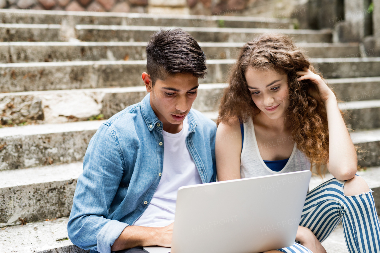 Attractive teenage student couple sitting on stone steps in front of university with laptop, reading or watching something.