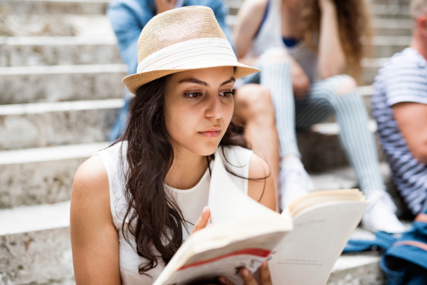 Attractive teenage student girl sitting on stone steps with her friends in front of university reading a book.