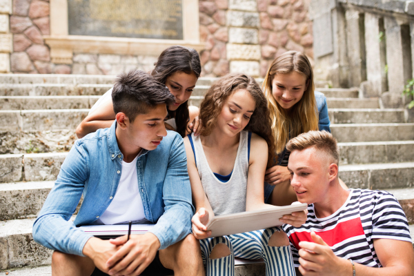 Group of attractive teenage students sitting on stone steps in front of university holding tablet, reading or watching something.